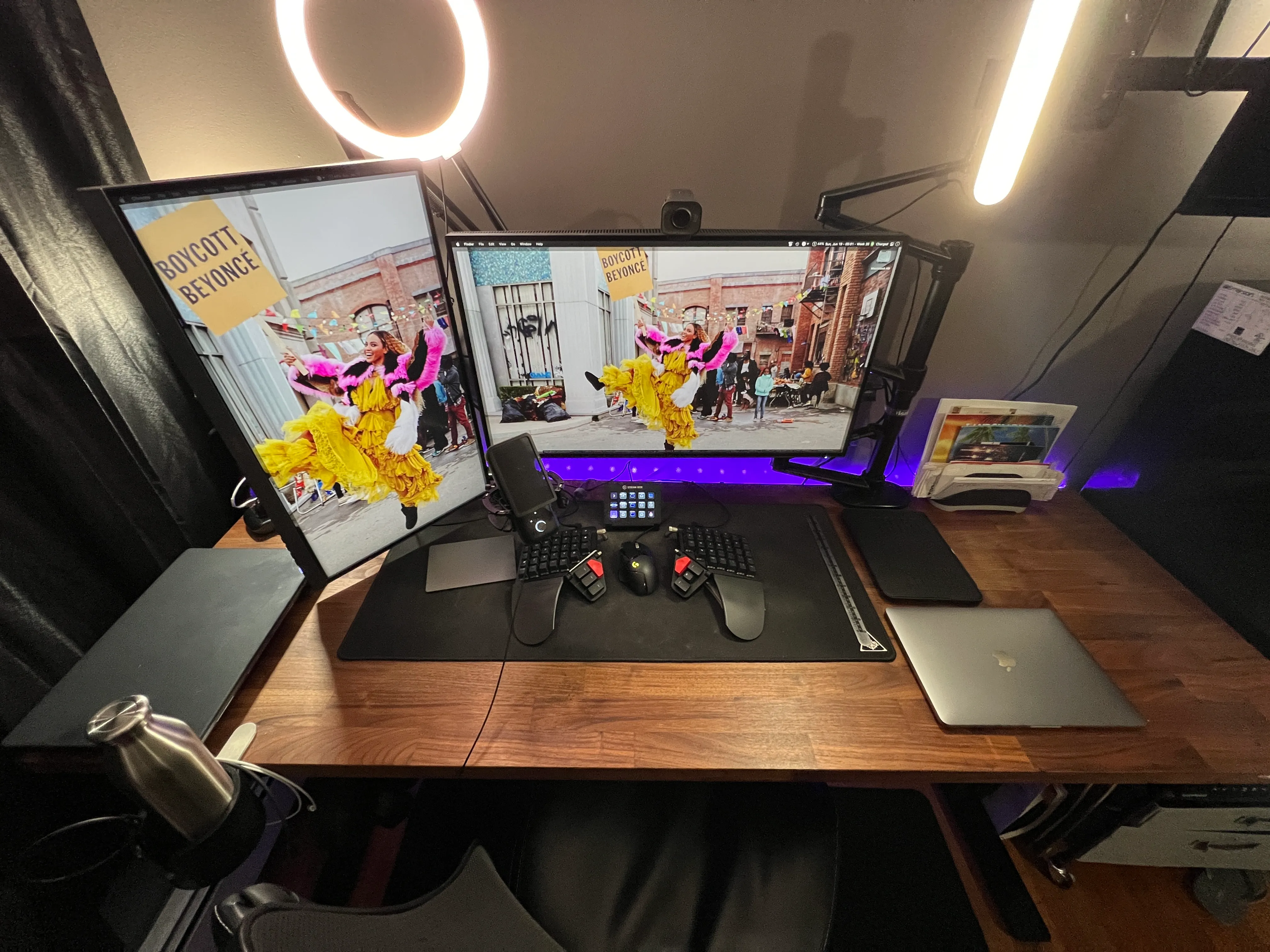Manny's work from home setup and gear. Featuring dual monitor displays: vertical to the left, horizontal to the right. A split keyboard with mouse in the center and track pad to the left. A stream deck, webcam and 2 lights. A personal and work laptop on opposite ends.